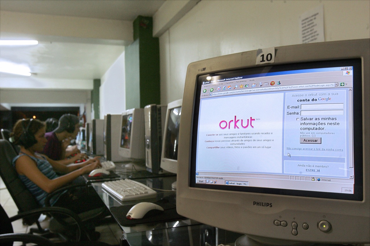 Tela de login do Orkut numa lan house, fileira de computadores com pessoas usando fones de ouvido ao fundo