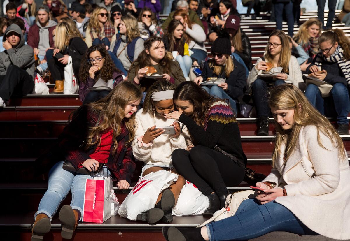 Grupo de jovens rindo, aglomeradas em volta de um celular numa arquibancada